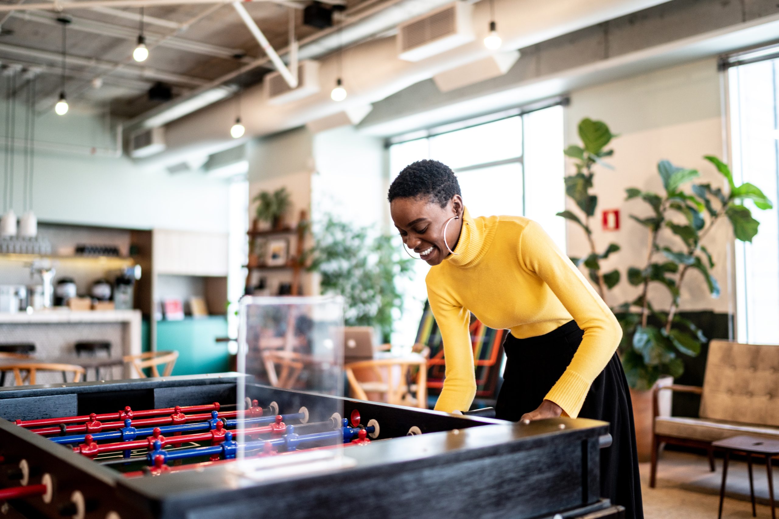 A woman in a yellow turtleneck and black skirt plays foosball in an office.
