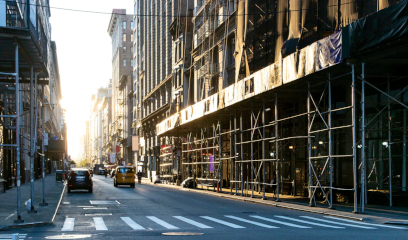 Construction scaffolding covers the buildings and sidewalks, cars passing by along 19th St. NYC