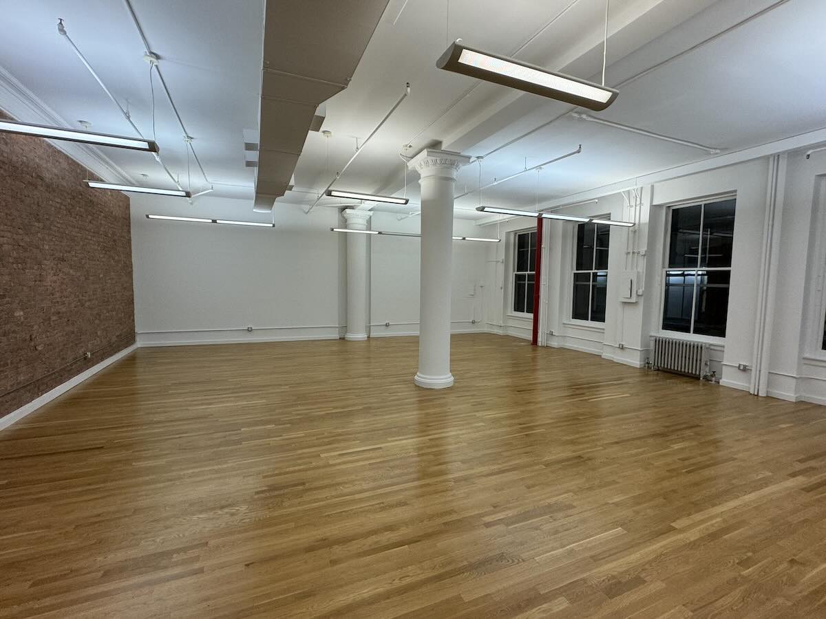 Empty room with polished floors, large windows, white walls, and brick wall.
