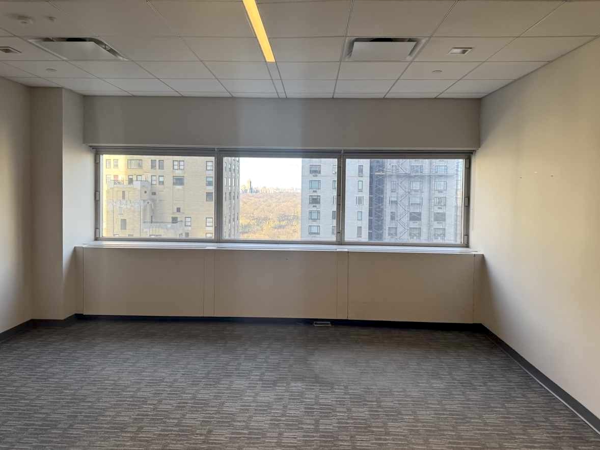 Empty office, carpeted, large window with cityscape view.