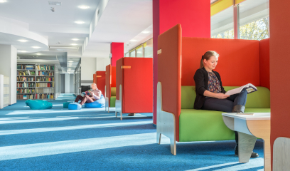 Woman reads in a bright library cubicle; bookshelves and windows nearby.