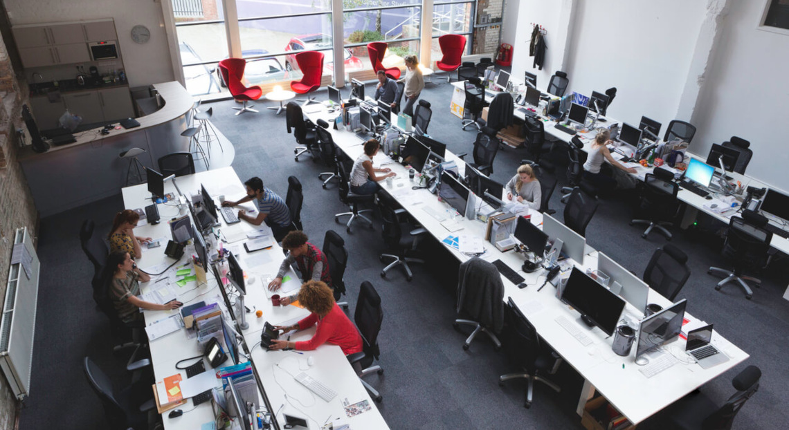 Modern office with people at desks, large windows, lounge, and kitchenette.